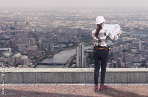 Female engineer holding construction plans standing on the roof top above a city © Ravil Sayfullin