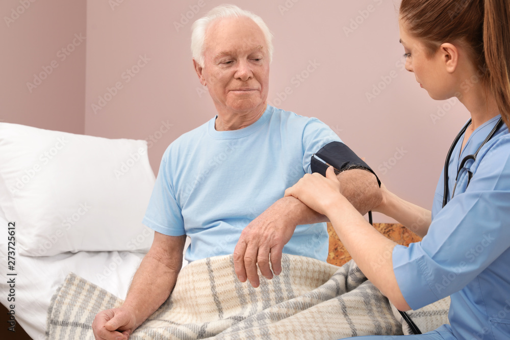 Fototapeta premium Nurse measuring senior man's blood pressure in hospital ward. Medical assisting