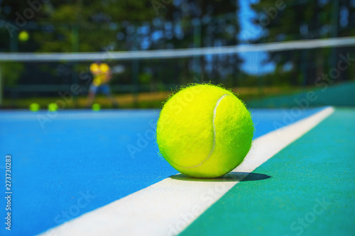 Tennis ball on white court line on hard modern blue green court with player, net, balls, trees on the background. Close-up, selective focus. Sport, tennis play, healthy lifestyle concept. © IrynaV