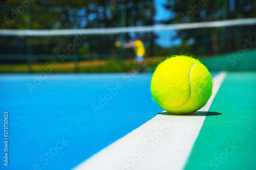 Tennis ball on white court line on hard modern blue green court with player, net, balls, trees on the background. Close-up, selective focus. Sport, tennis play, healthy lifestyle concept. © IrynaV