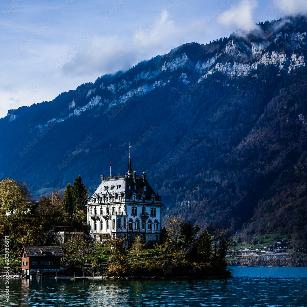View across Brienzersee Lake, Switzerland with the white facade of Iseltwald castle reflected in the calm waters of the lake and Niesen mountain rising up in the far distance