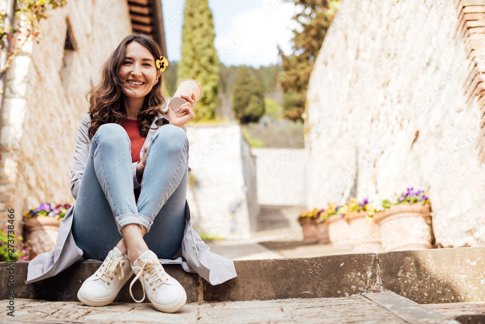 Beautiful smiling brunette woman sitting on the ground with a cup of coffee to go - relaxing in the center of a European (Italian) medieval town