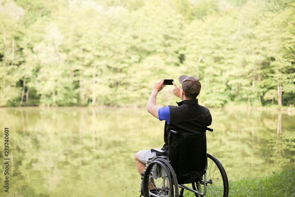 Happy and young disabled or handicapped man sitting on a wheelchair in nature. Photographing landscape with smartphone.