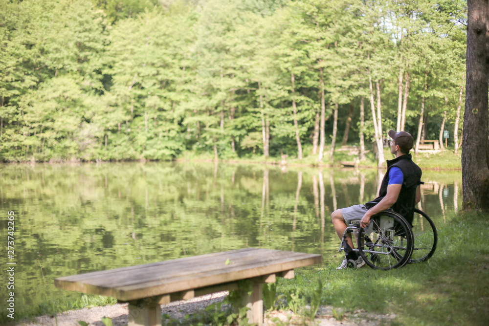 Happy and young disabled or handicapped man sitting on a wheelchair looking at beautiful lake in nature.