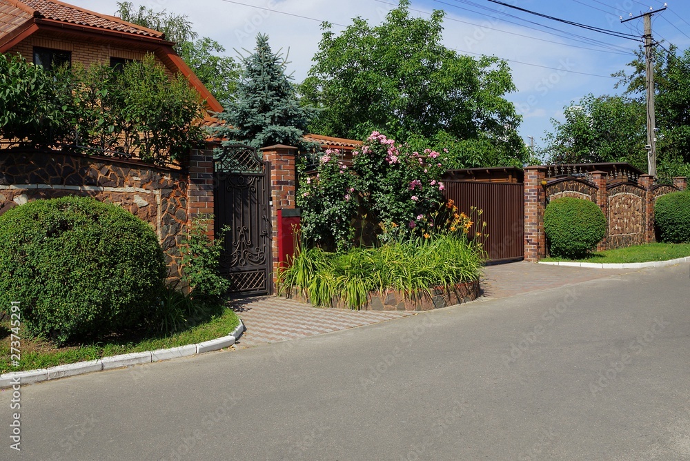 brown fence and gate on the street with green vegetation and flower bed along the asphalt road brown fence and gate on the street with green vegetation and flower bed along the asphalt road