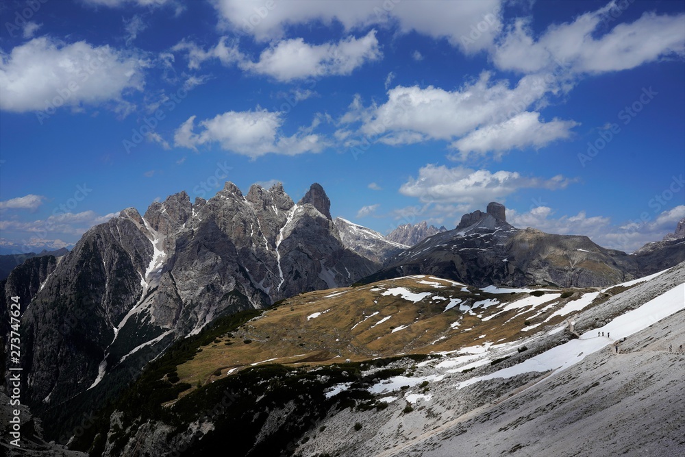 Mountain scenery, peaks and snow on the ground. A small path on the side of the mountain and people walking on it. Dolomites, Italy, June 