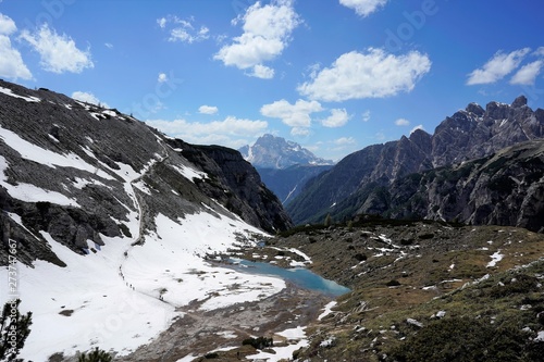 Landscape in the mountains, seen from above, with a path with people walking on it, a small lake and snow. Dolomites, Italy, June 