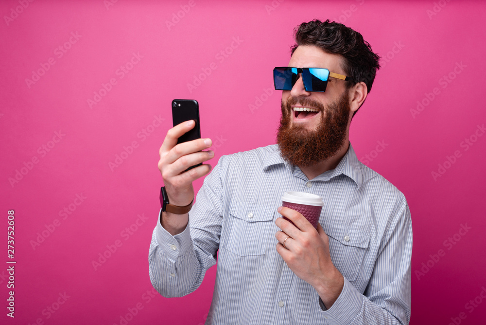 Young bearded hipster man taking a selfie with his cup of coffee on pink background. Young bearded hipster man taking a selfie with his cup of coffee on pink background.