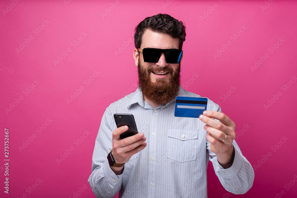Bearded man enjoying online shopping or internet banking holding his phone and credit card on pink background. Bearded man enjoying online shopping or internet banking holding his phone and credit card on pink background.