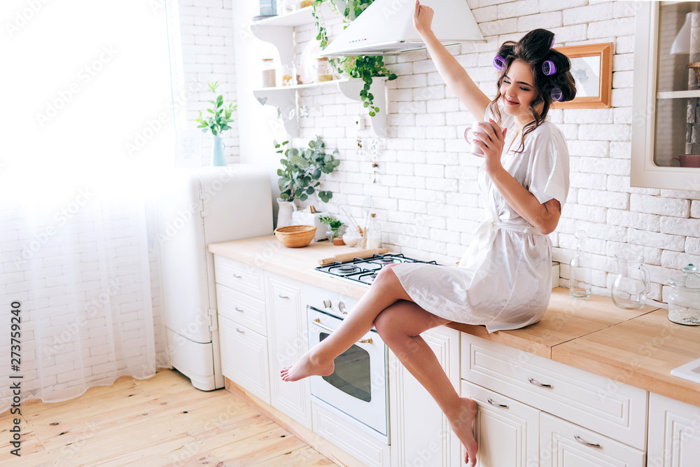 Young careless housewife stretching hand up and enjoying life. Hold cup in hand. Curlers in hair. Alone in kitchen. Sugar daddy pays for everything. Wearing white dressing gown. Young careless housewife stretching hand up and enjoying life. Hold cup in hand. Curlers in hair. Alone in kitchen. Sugar daddy pays for everything. Wearing white dressing gown.