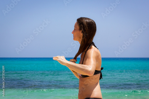 Woman in black bikini doing wet hair by turquoise water sea. Female tourist by shore on the beach after swimming. Summertime, relax vacation, travel destination concepts