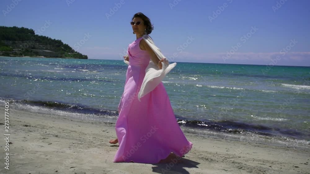 Portrait of charming brunette woman wearing pink dress and white shawl on her shoulder taking a happy walk on beach shore. Portrait of charming brunette woman wearing pink dress and white shawl on her shoulder taking a happy walk on beach shore.