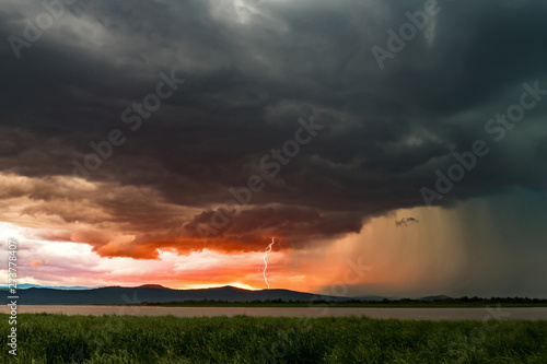 Beautiful fiery clouds during sunset and scary thunderclouds with thunder lighthing striking the ground. Dramatic thunderstorm scene.