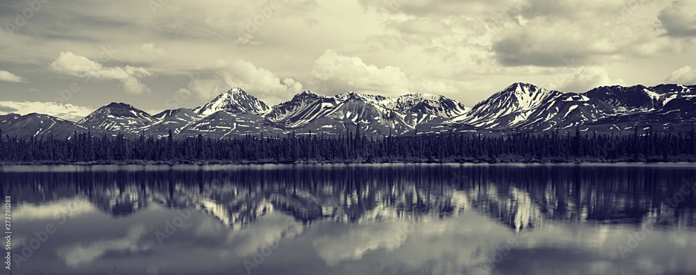 Jagged snowy mountain peaks with snow on the top reflected in shallow river during early summer. 