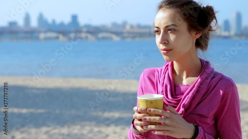 portrait of beautiful young athletic woman in pink sportswear hoodei drinking coffee from paper cup on the city sand beach at sunny morning. sea and city on the background. photo