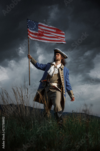 Stampa su Tela American revolution war soldier with flag of colonies over dramatic landscape