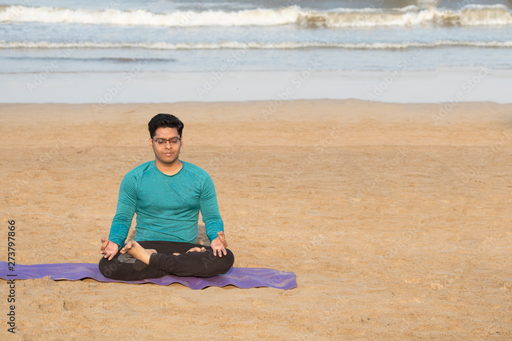 Fototapeta premium Young man meditating in yoga lotus position at serene beach in morning sunlight