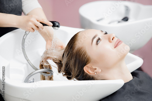 Concept beauty salon. Hairdresser washes hair of beautiful blonde girl under tap in wash