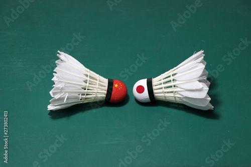 Two used shuttlecocks on green floor of Badminton court with both head each other. One head painted with Chinese flag And one head painted with the Japanese flag.