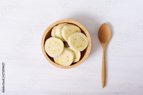 Sliced banana in a bowl and wooden spoon on white background, top view