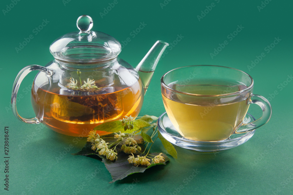 Glass tea cup and pot filled with linden tea and linden stem on green background. Side view Glass tea cup and pot filled with linden tea and linden stem on green background. Side view