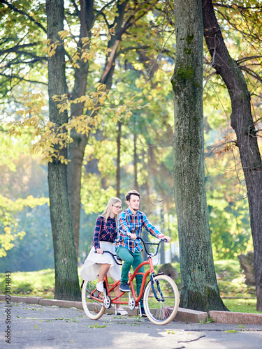 Young active couple, bearded man and attractive long-haired blond woman riding tandem bicycle along asphalt path in lit by bright sun beautiful park under tall trees with green and yellow leaves.