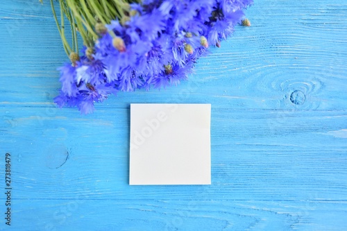 card for invitation or congratulation with bouquet of blue summer flowers. Bunch of beautiful cornflower on turquoise wooden table with empty paper for text. Bluet flower with blank note. Copy space