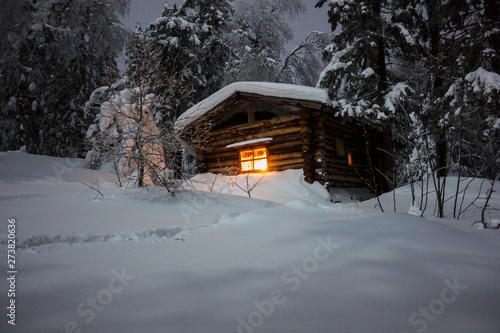 The light of a Christmas candle in the window of a lonely wooden log hut in a night winter dense forest under white fluffy snow