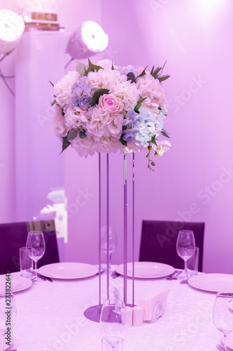 Beautiful table setting with crockery and white flower arrangement in a vase on a high stem for a party, wedding reception or other festive event. Glassware and cutlery for catered event dinner © Sergei Shavin