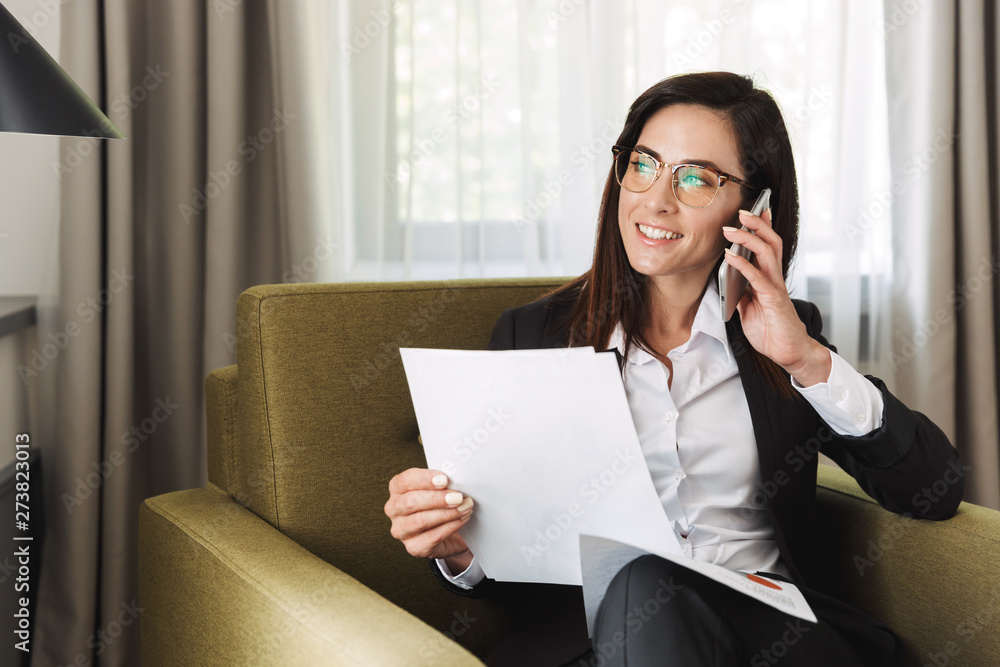 Young business woman in formal wear clothes indoors at home talking by mobile phone work with documents. Young business woman in formal wear clothes indoors at home talking by mobile phone work with documents.
