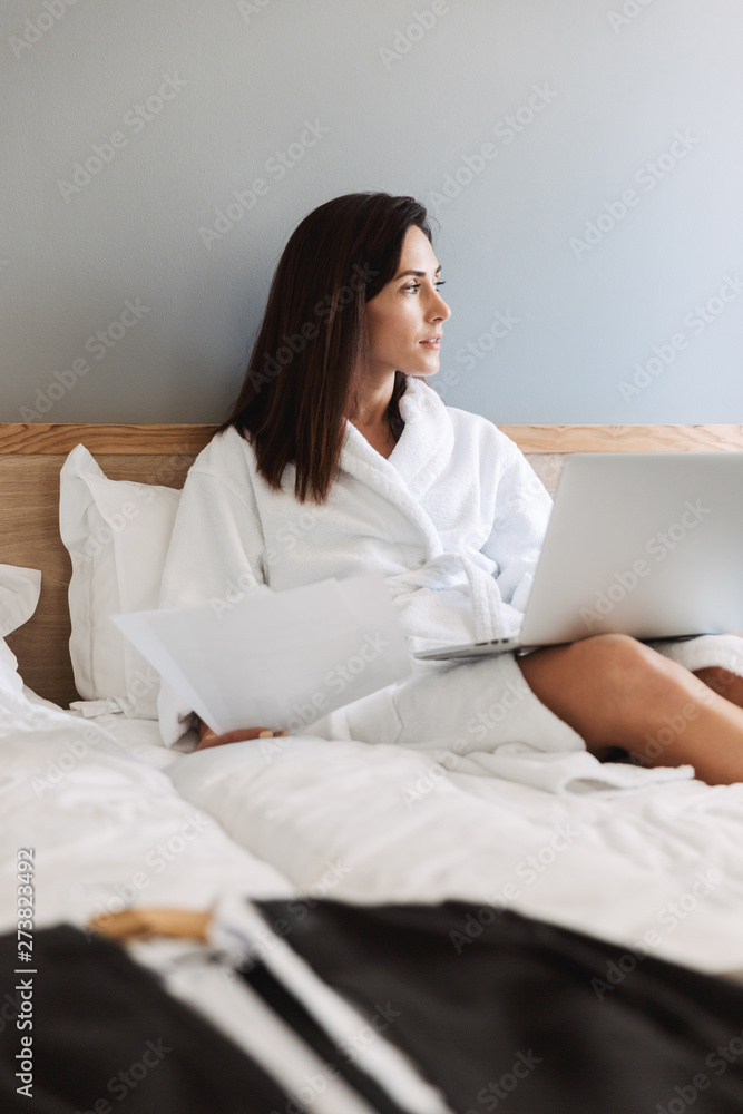 Portrait of beautiful adult businesswoman working with paper documents and laptop while lying on bed in apartment Portrait of beautiful adult businesswoman working with paper documents and laptop while lying on bed in apartment