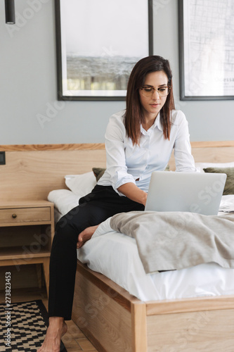 Portrait of beautiful middle-aged businesswoman working on laptop while sitting on bed in apartment