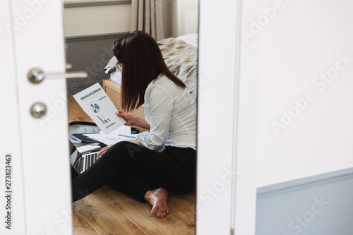 Portrait of brunette adult businesswoman working with paper documents and laptop while sitting on floor near luggage in apartment