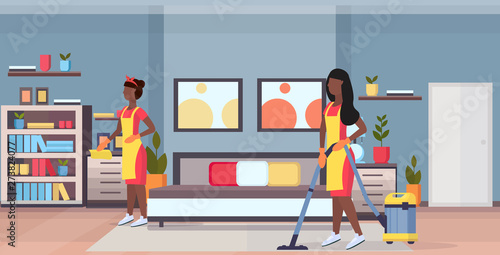 janitors women cleaners in uniform working together african american girls using vacuum cleaner and dust cloth cleaning service concept modern bedroom interior flat full length horizontal