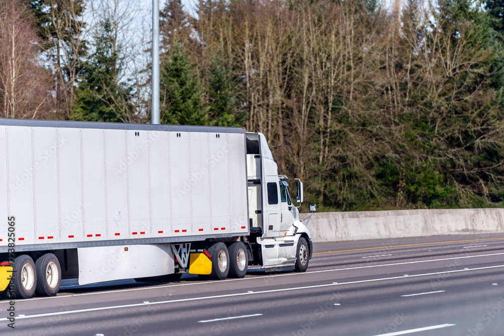 Big rig white semi truck transporting commercial cargo in dry van semi trailer driving on the straight wide highway road