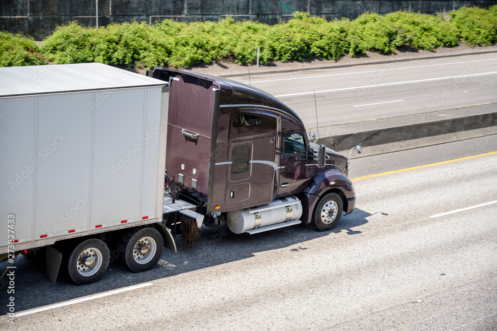 Dark brown big rig commercial long haul semi truck transporting dry van semi trailer on the divided highway road
