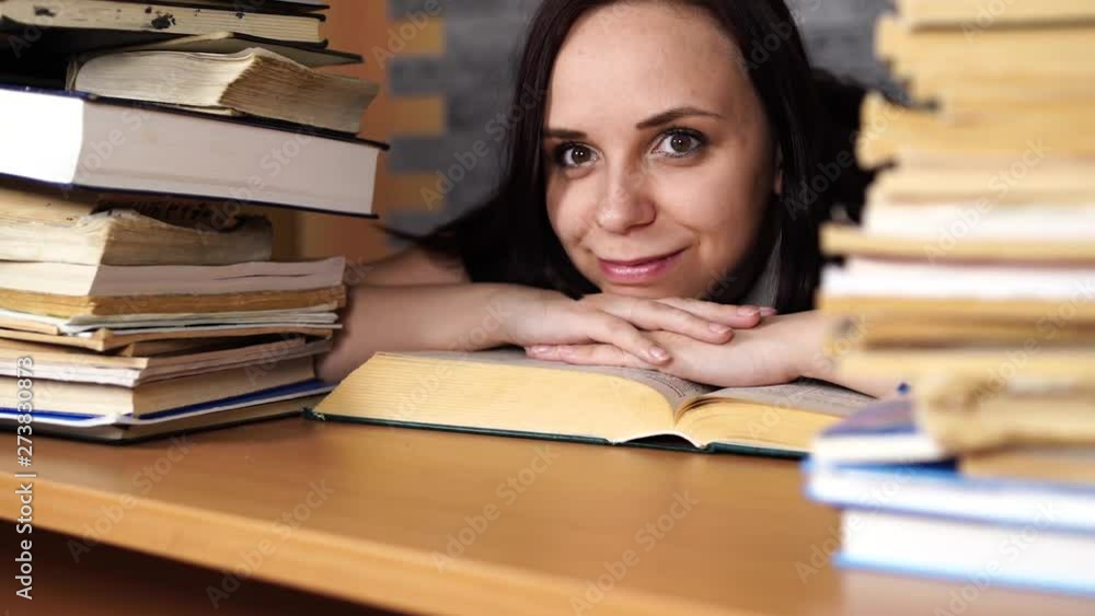 Tired female student reading among books. Pensive young woman sitting at table with pile of book and reading on background of grey wall Tired female student reading among books. Pensive young woman sitting at table with pile of book and reading on background of grey wall
