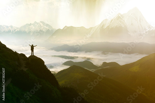 Wide mountain panorama. Small silhouette of tourist with backpack on rocky mountain slope with raised hands over valley covered with white puffy clouds. Beauty of nature, tourism and traveling concept