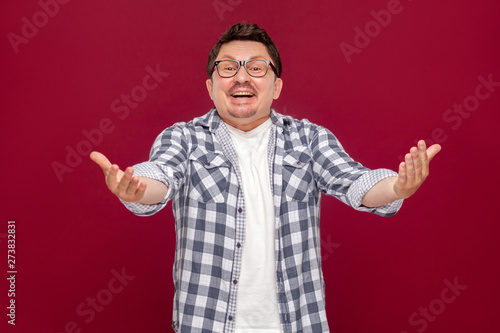 Portrait of excited handsome middle aged business man in casual checkered shirt, eyeglasses standing, toothy smile and looking at camera with raised arms. studio shot, isolated on dark red background