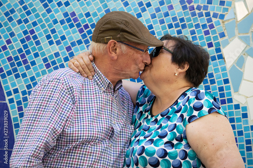 Pareja de personas mayores (abuelos) enamoradas, besándose delante de un mosaico azul en un parque urbano. photo
