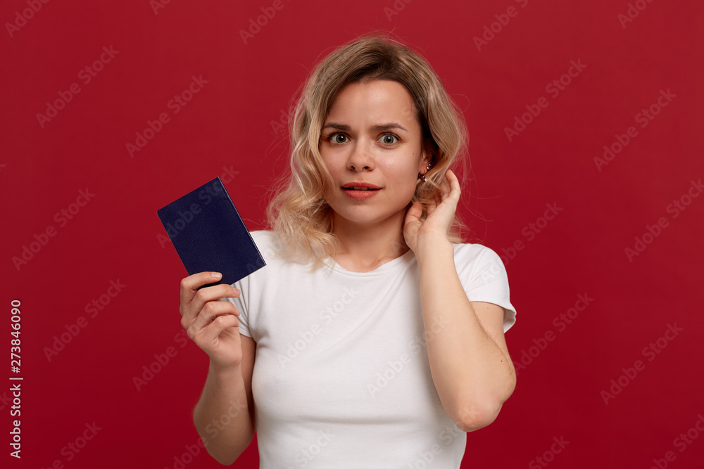 Naklejka premium Portrait of a beautiful girl with curly blond hair dressed in a white t-shirt standing on a red background. Model smiles at the camera and holds passport of blue color.