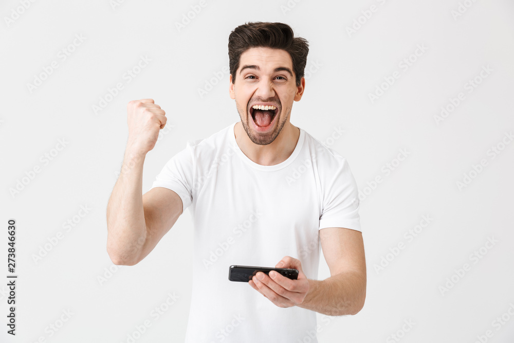 Excited happy emotional young man posing isolated over white wall background play games by mobile phone make winner gesture. Excited happy emotional young man posing isolated over white wall background play games by mobile phone make winner gesture.