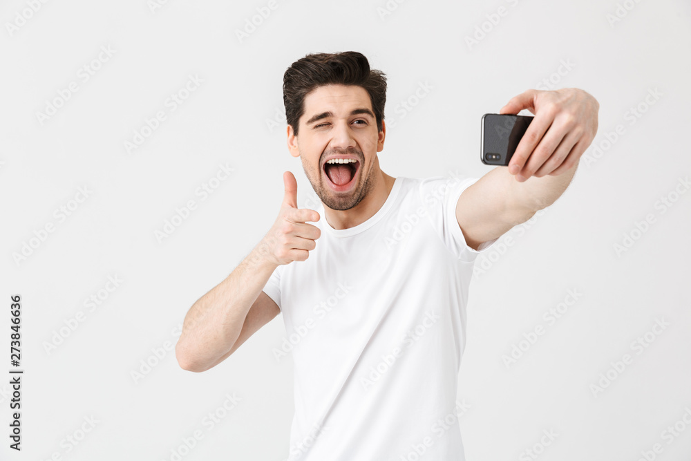 Emotional young man posing isolated over white wall background pointing take selfie by mobile phone. Emotional young man posing isolated over white wall background pointing take selfie by mobile phone.