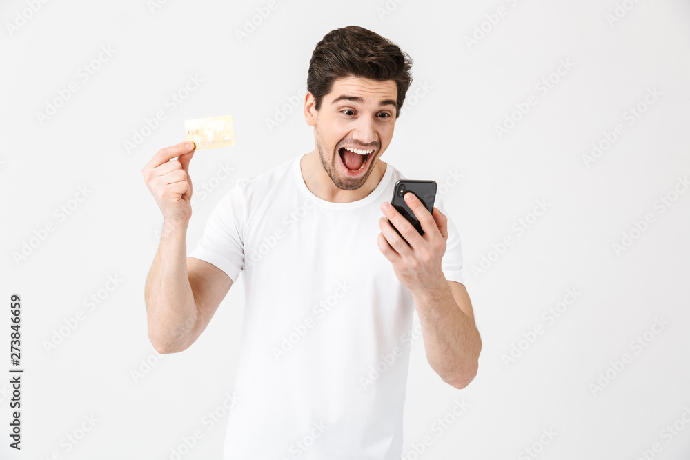 Excited happy young man posing isolated over white wall background using mobile phone holding credit card. Excited happy young man posing isolated over white wall background using mobile phone holding credit card.