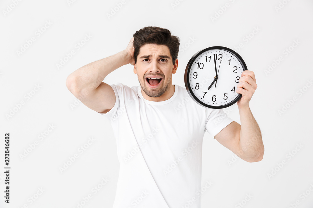 Screaming confused young man posing isolated over white wall background holding clock. Screaming confused young man posing isolated over white wall background holding clock.