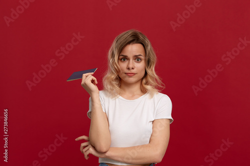 Portrait of a beautiful girl with curly blond hair dressed in a white t-shirt standing on a red background and holding passport.