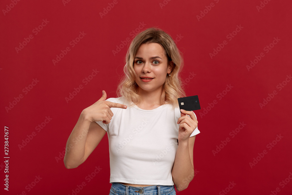 Portrait of a beautiful girl with curly blond hair dressed in a white t-shirt standing on a red background. Model holds bank card and points at it with finger Portrait of a beautiful girl with curly blond hair dressed in a white t-shirt standing on a red background. Model holds bank card and points at it with finger