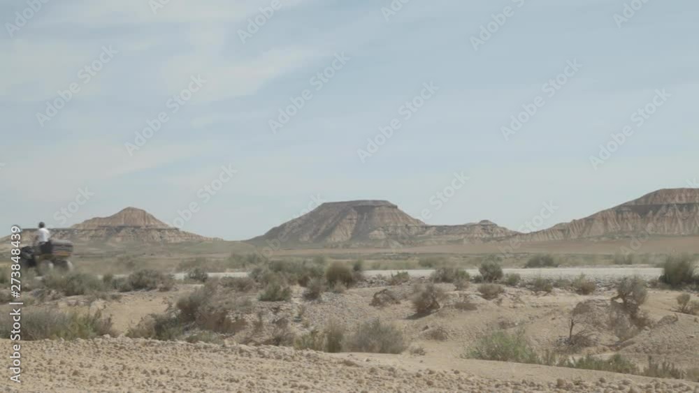 Driver man in white shirt standing and driving on his quad and making dust on the ground with three huge mountains in the background in empty desert landscape around an isolated wild road. Driver man in white shirt standing and driving on his quad and making dust on the ground with three huge mountains in the background in empty desert landscape around an isolated wild road.