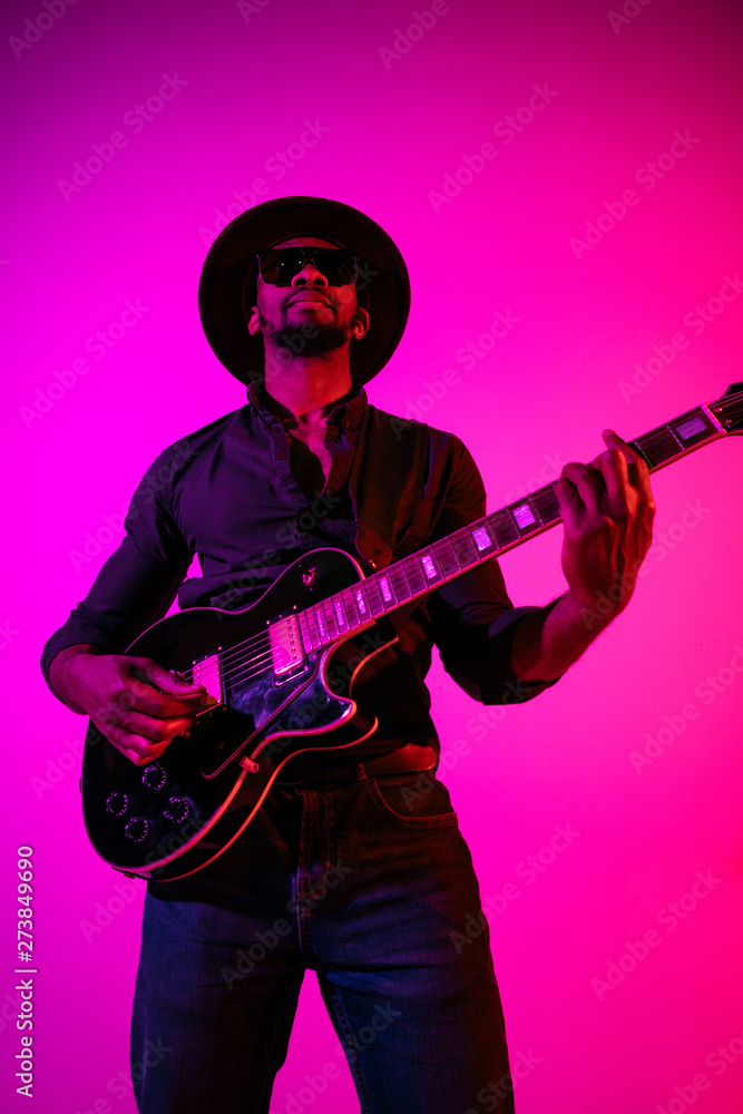 Obraz premium Young african-american musician playing the guitar like a rockstar on gradient purple-pink background in neon light. Concept of music, hobby. Joyful attractive guy improvising. Colorful portrait.