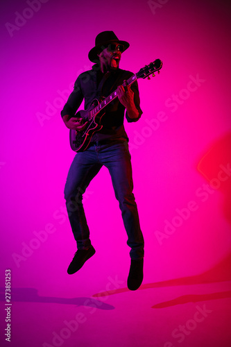 Young african-american musician playing the guitar like a rockstar on gradient purple-pink background in neon light. Concept of music, hobby. Joyful attractive guy improvising. Colorful portrait.